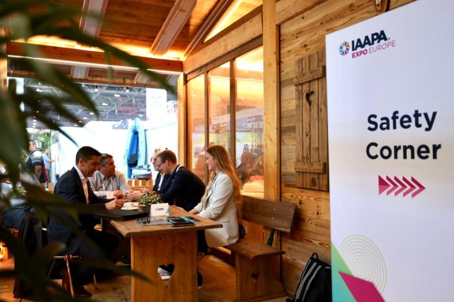 A banner in IAAPA corporate branding with "Safety Corner" label in camera focus, with an arrow pointing offscreen. Background shot, multiple people sitting together in a group talking at IAAPA EMEA Safety meeting